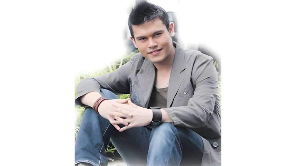 Vince Chong sitting with his arms resting on his knees, smiling towards the camera, wearing a grey coat and denim jeans