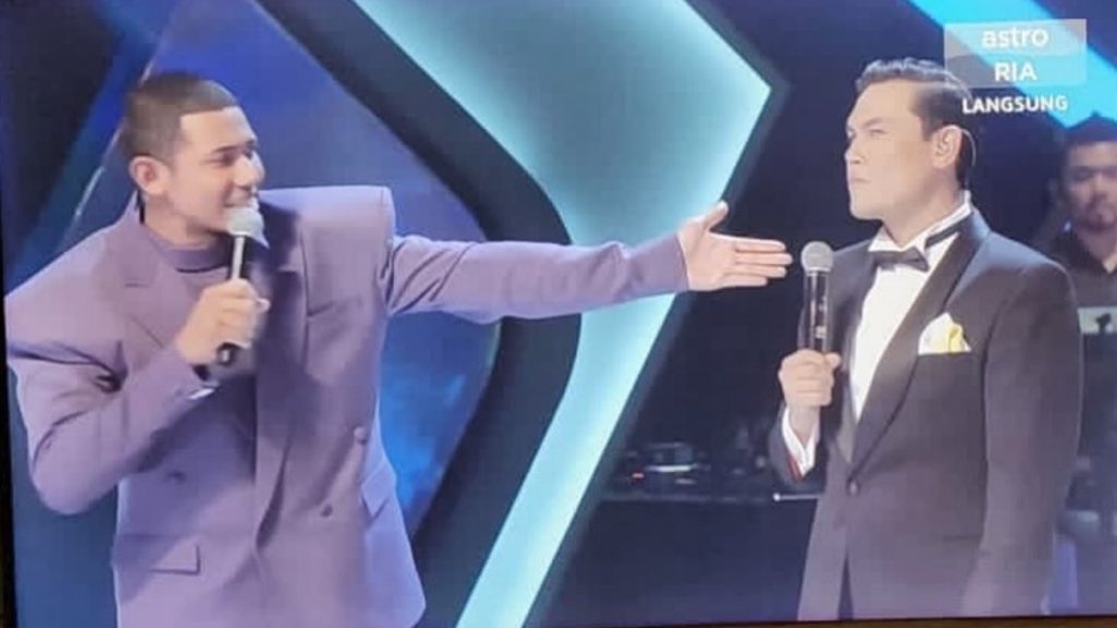 Two men onstage (Vince Chong on the right) wearing suits, with a blue backdrop