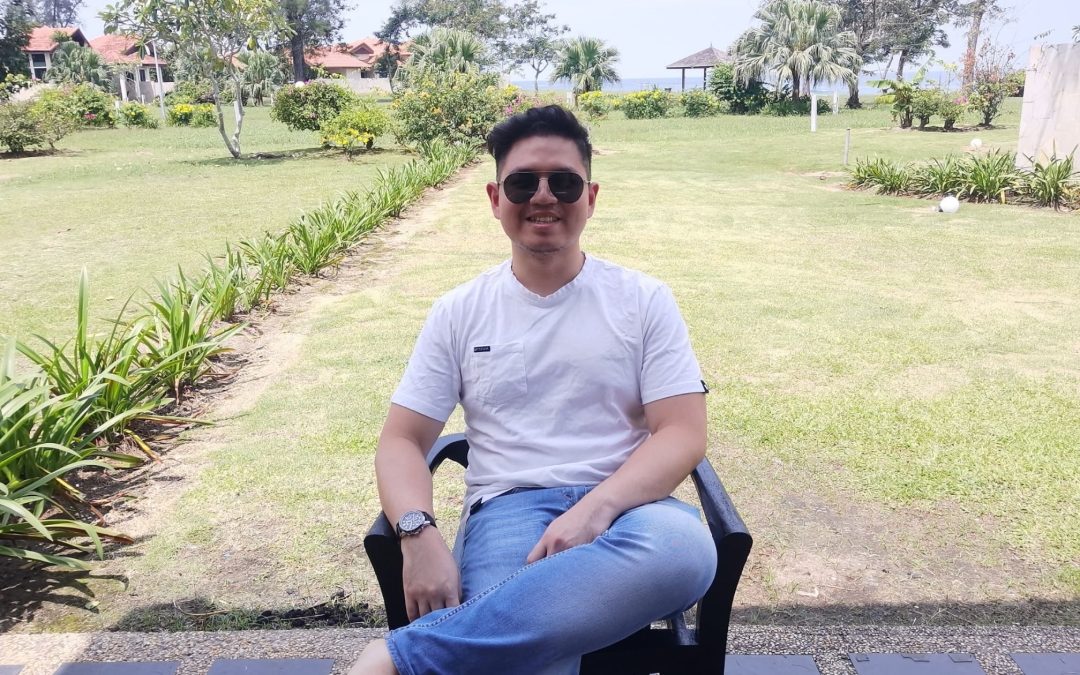 Young man in his 20s, wearing a white shirt with denim jeans, sitting on a chair in a garden with a beach far off